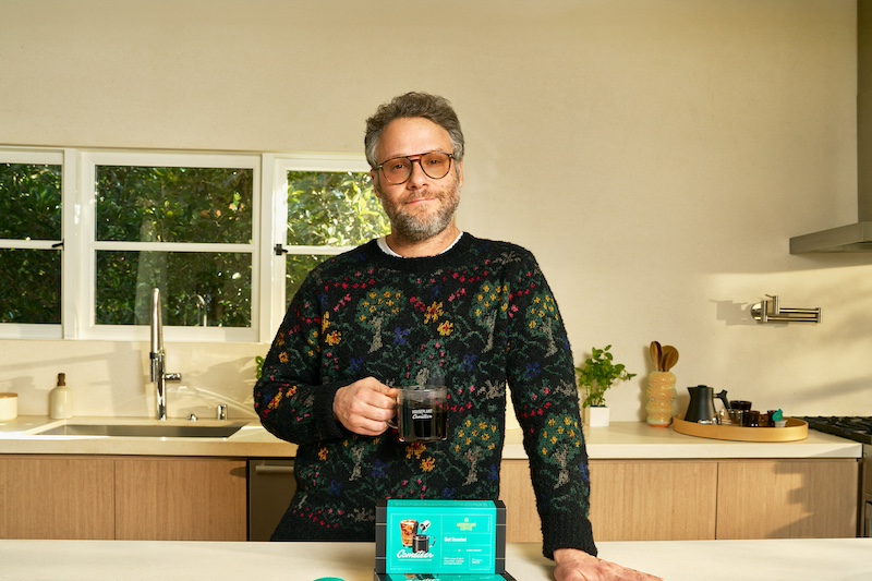 Seth Rogen in a floral sweater drinking a hot mug of coffee in a kitchen.